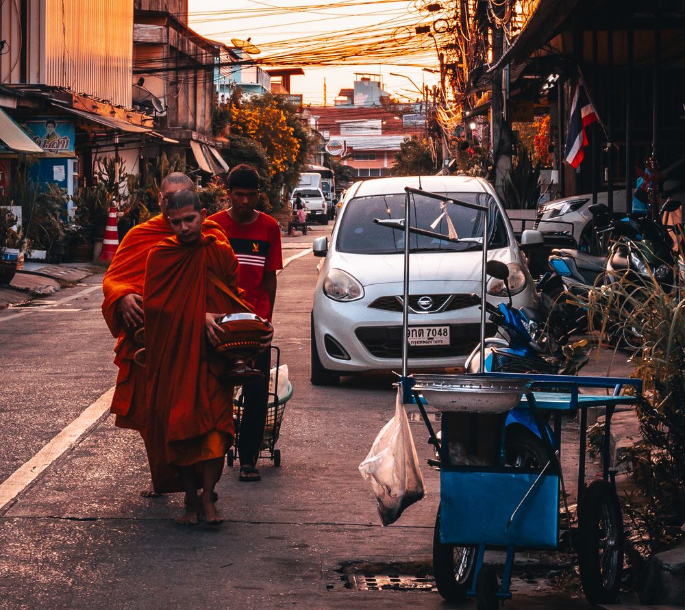 Busy street scene in Thailand with robed figures walking past parked cars during golden sunset hour.