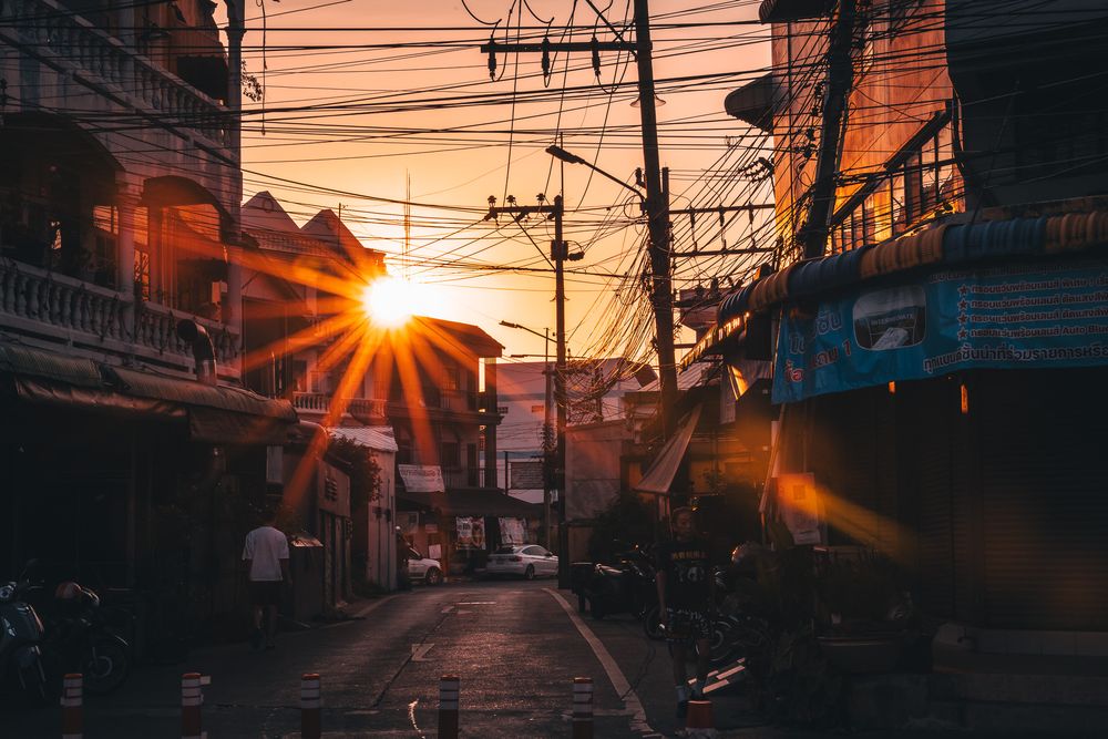 Sunset through power lines illuminates a narrow urban street in an Asian city with warm orange light.