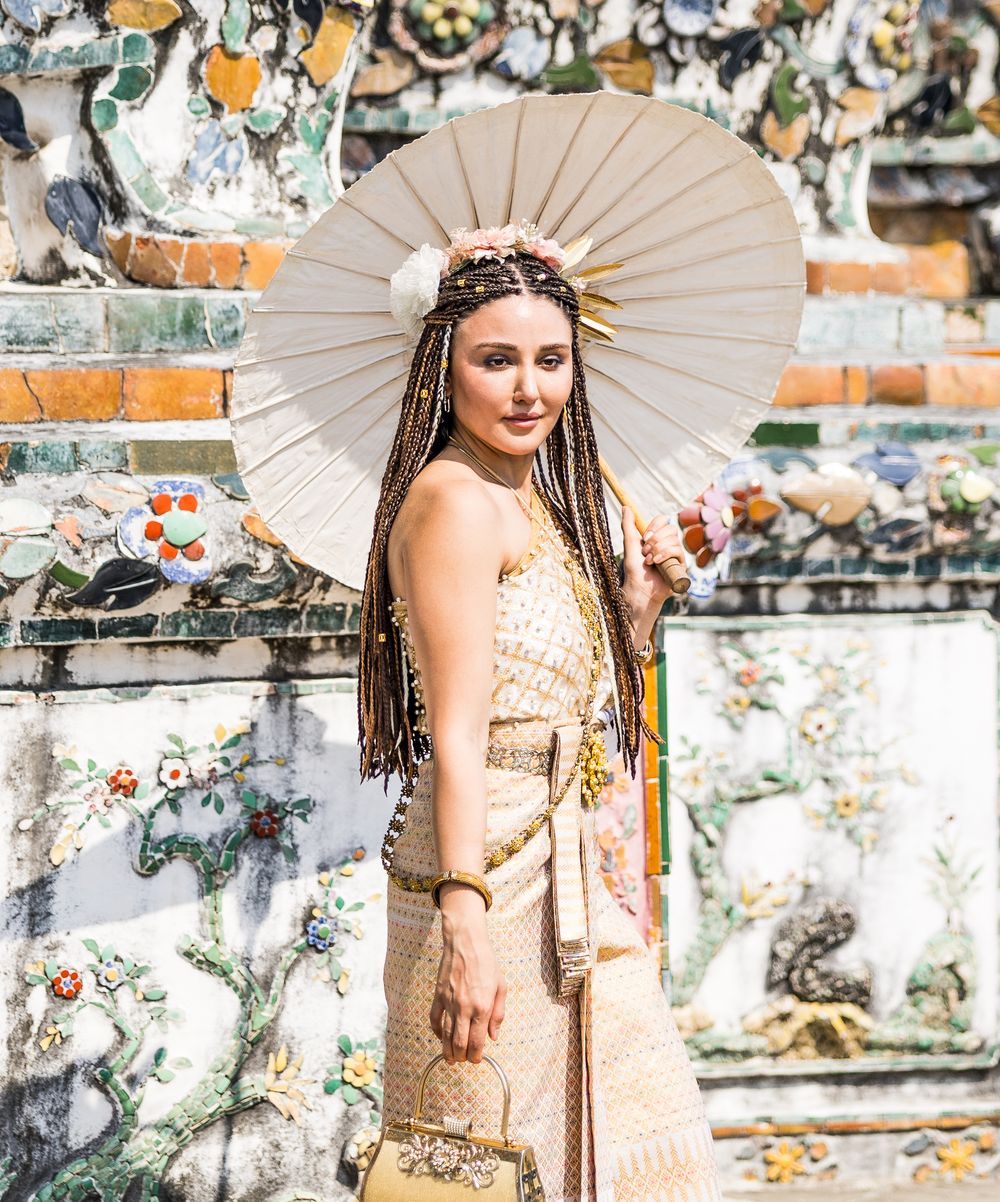A figure in a boho-style dress and headpiece poses with a parasol against an ornate temple wall in Thailand.