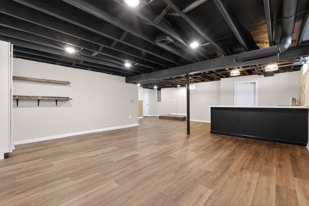 Finished basement recreation room with light hardwood floors, black bar counter, and exposed ceiling.