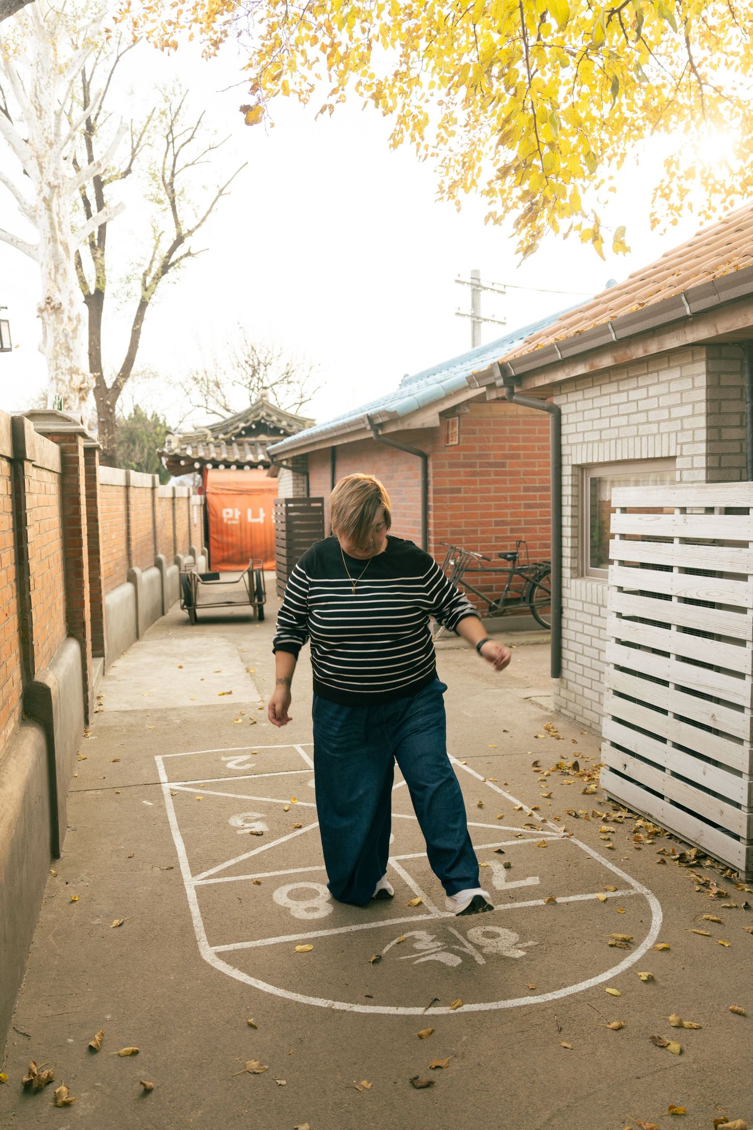 Person in striped shirt plays hopscotch on concrete path with autumn leaves falling from trees in background.