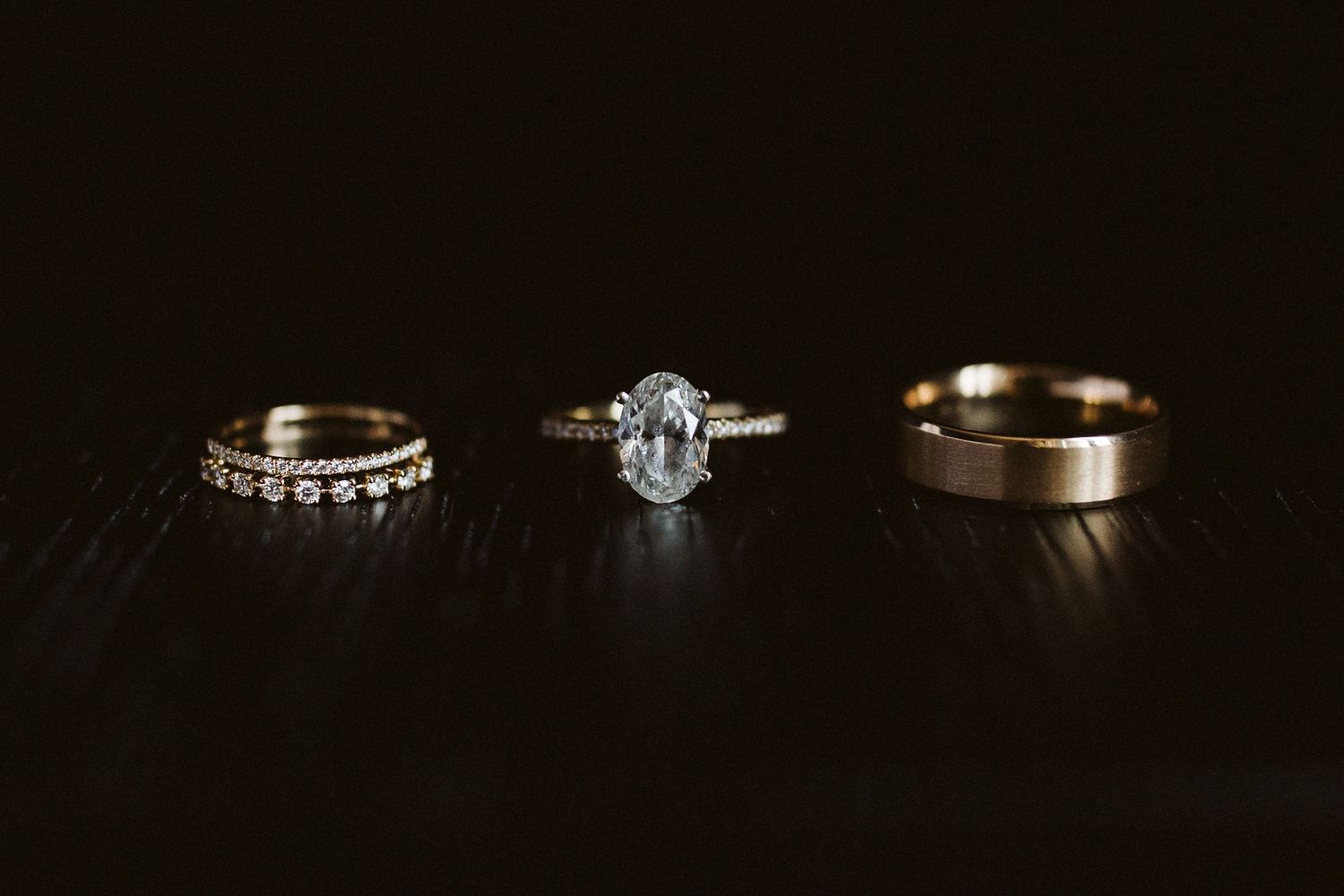 Three wedding rings photographed on dark wood surface including engagement ring and bands.