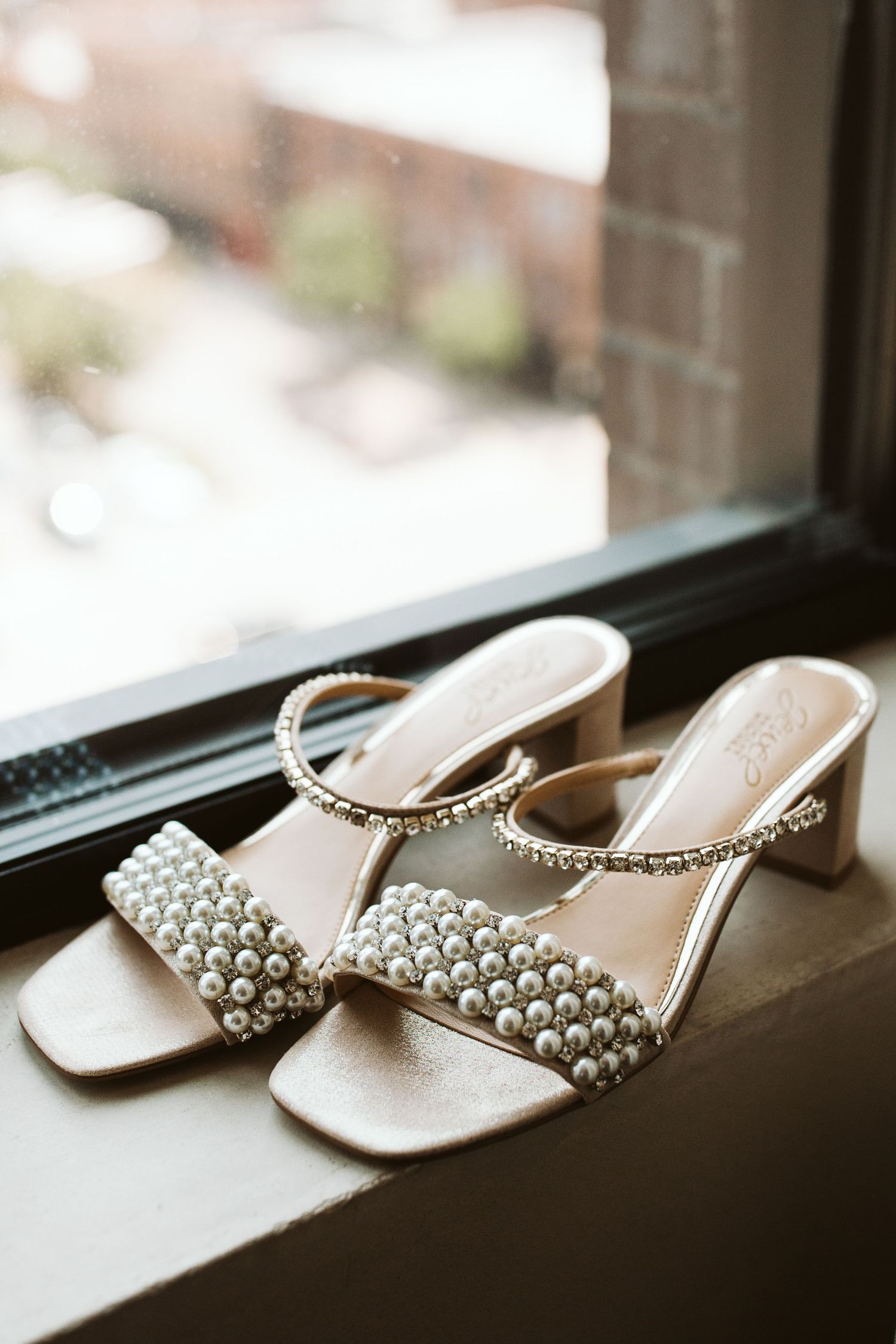 Elegant pearl-embellished wedding sandals with crystal straps sitting on a windowsill.