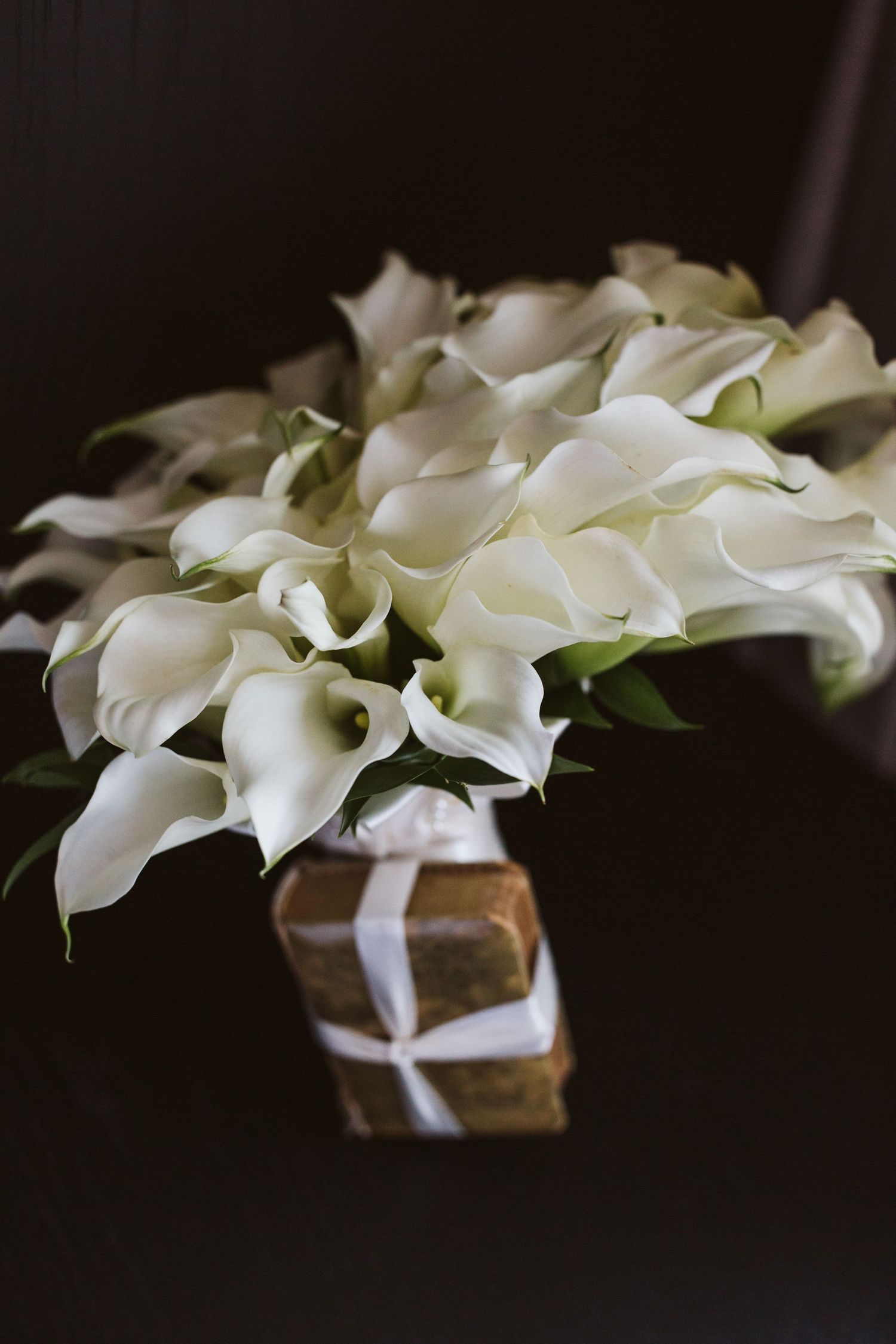 Elegant white calla lily bouquet arranged in a wrapped vase against dark background.