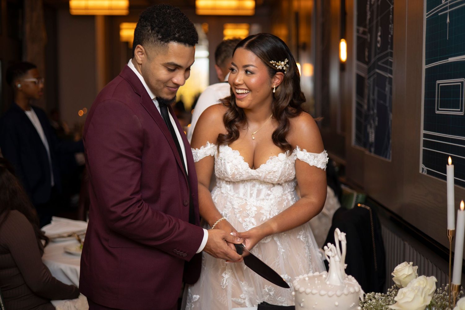 A joyful couple cuts their wedding cake together at their reception, with the bride in white and groom in burgundy.