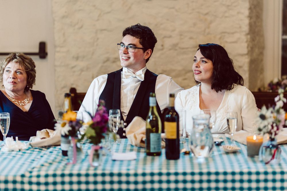 Wedding reception guests seated at table with checkered tablecloth, wine bottles and floral centerpieces.
