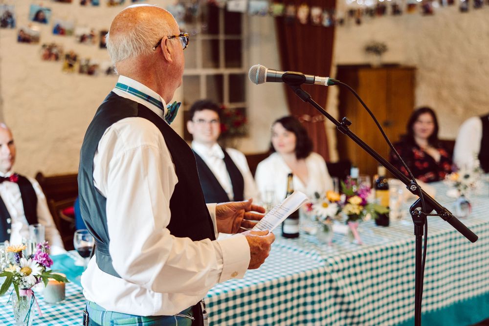 A speaker in formal attire delivers remarks at a banquet with a gingham tablecloth and floral centerpieces.