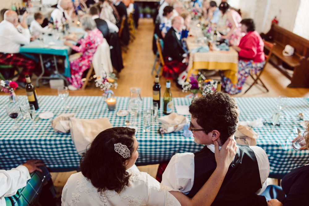 Wedding reception guests seated at long tables with blue checkered tablecloths in a community hall.