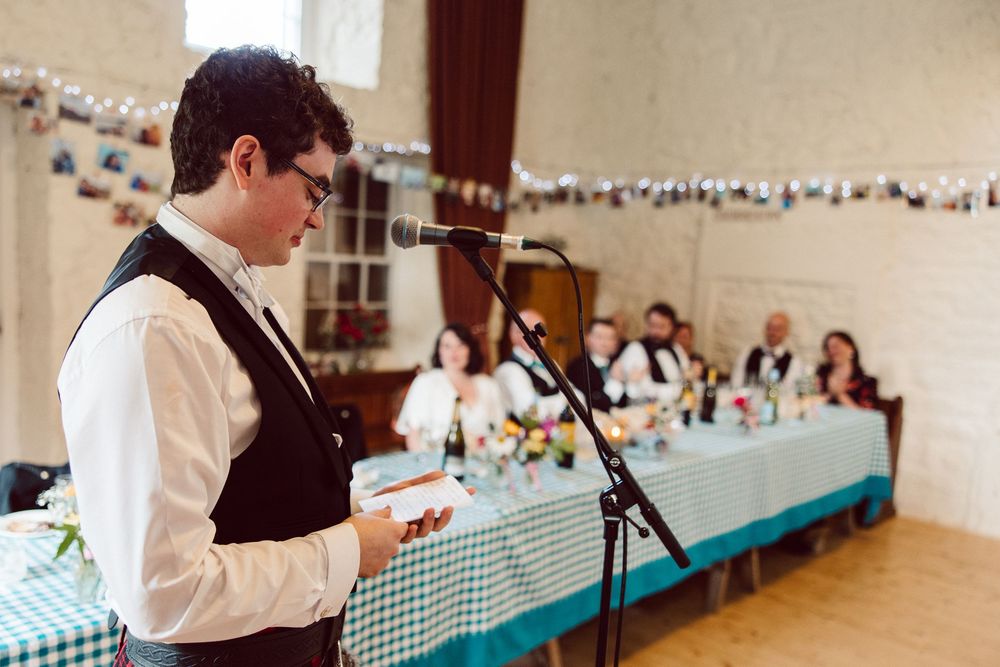 Speaker stands at microphone with string lights and blue checkered tablecloths in background at wedding reception.