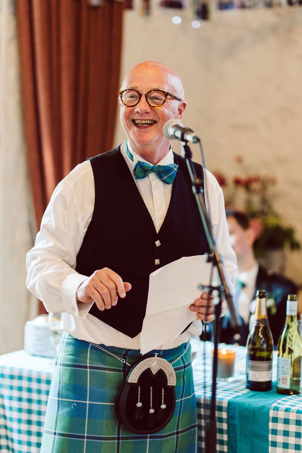 A speaker in a kilt, bow tie and vest smiles while standing at a microphone during a celebration with checkered tablecloths.
