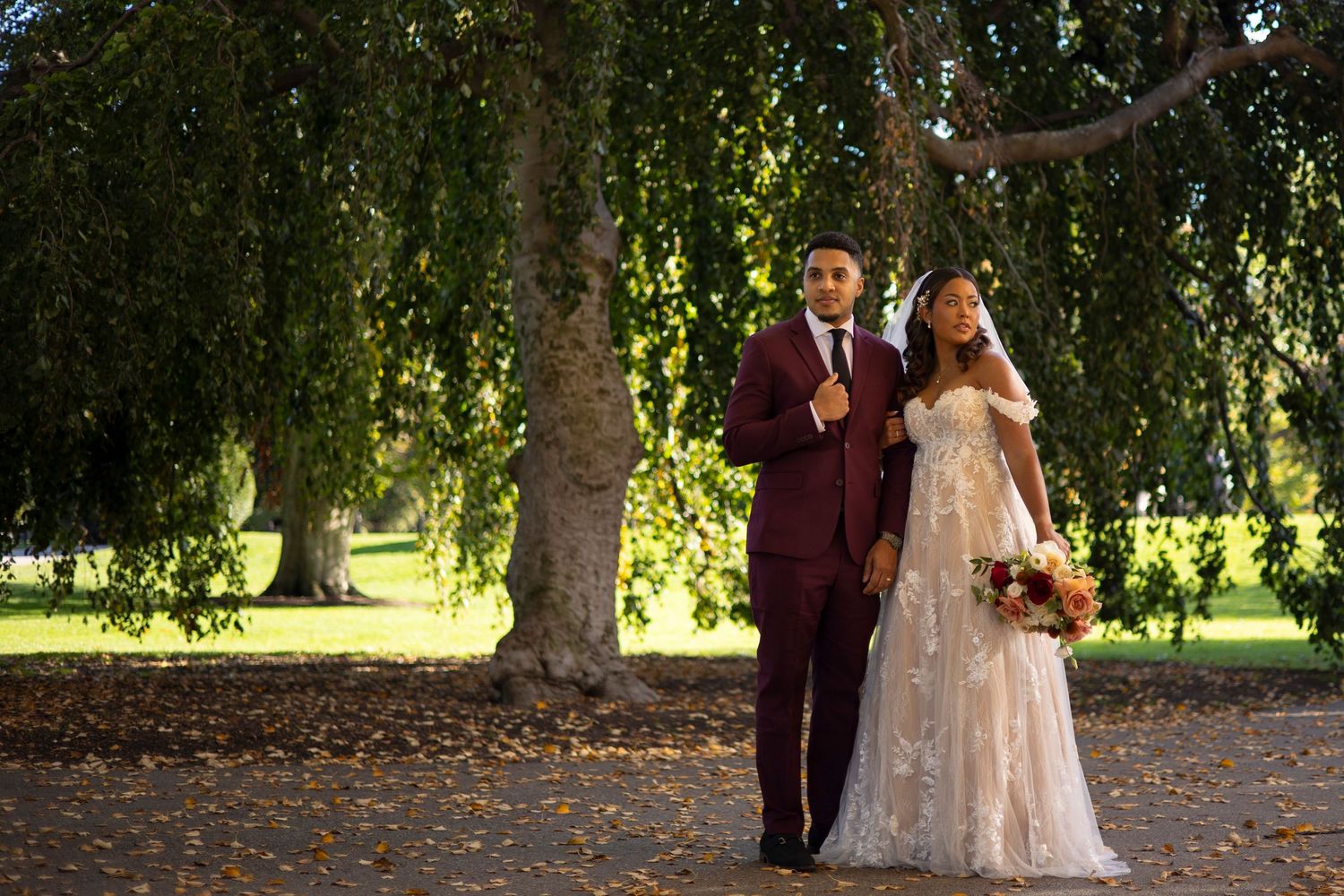A couple in wedding attire poses beneath autumn trees in a park during their outdoor photo session.
