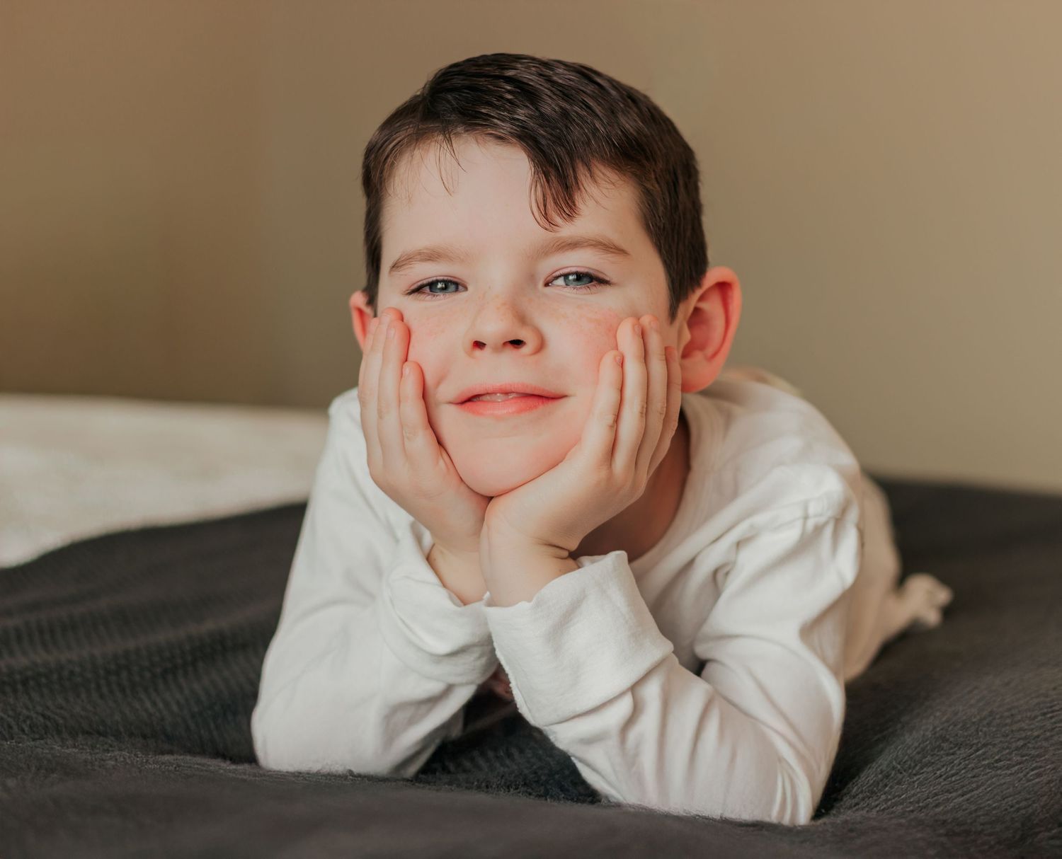 A young boy rests his chin on his hands, smiling warmly while lying on a gray bed | Westminster, CO motherhood & family photographer