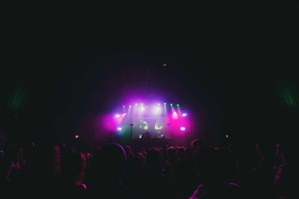 Concert crowd silhouettes against dark stage with vibrant purple and pink lighting effects during live performance.