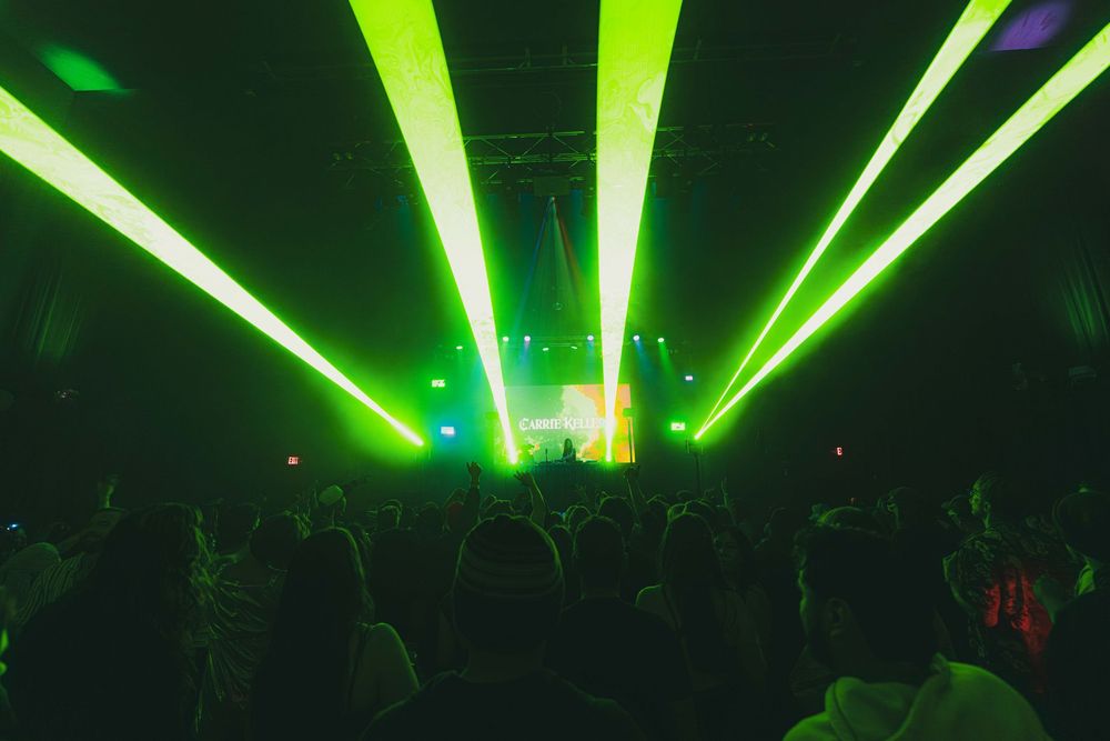 Bright green laser beams illuminate a dark concert venue as silhouetted crowd watches the performance.