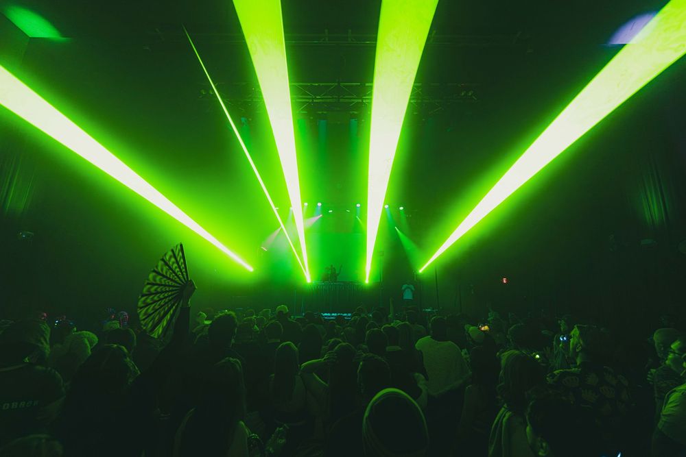 Bright green laser beams illuminate a dark nightclub with silhouetted crowd during an electronic music performance.