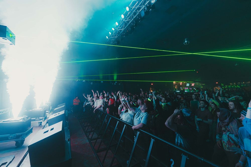 Concert crowd enjoys performance with bright turquoise lighting and laser beams in a smoky indoor venue.
