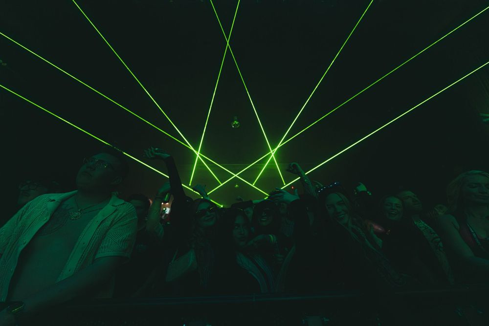 Green laser beams illuminate a dark concert venue as silhouetted crowd watches the performance.