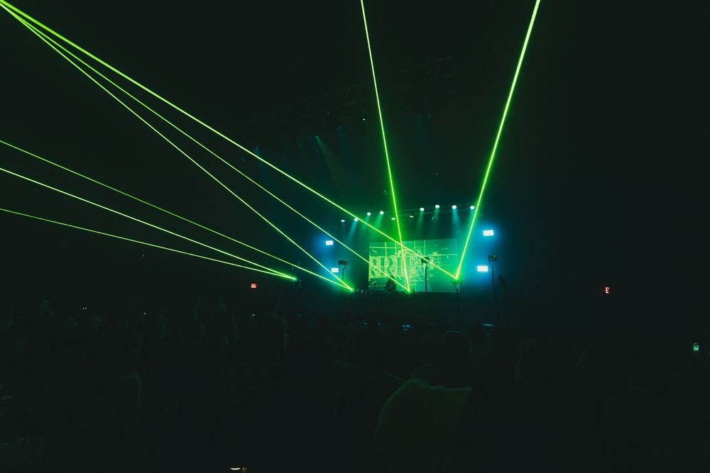Bright green laser beams illuminate a dark concert stage with blue lighting effects and fog.