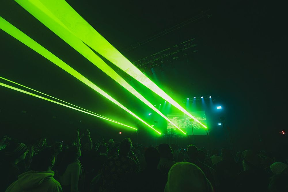 Bright green laser beams illuminate a dark concert venue as crowd silhouettes watch the stage performance.