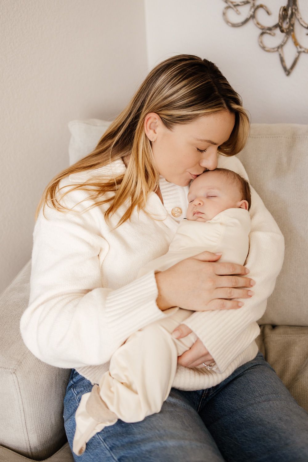 Mother in white sweater cuddles with baby in cream outfit on light gray couch in tender moment.