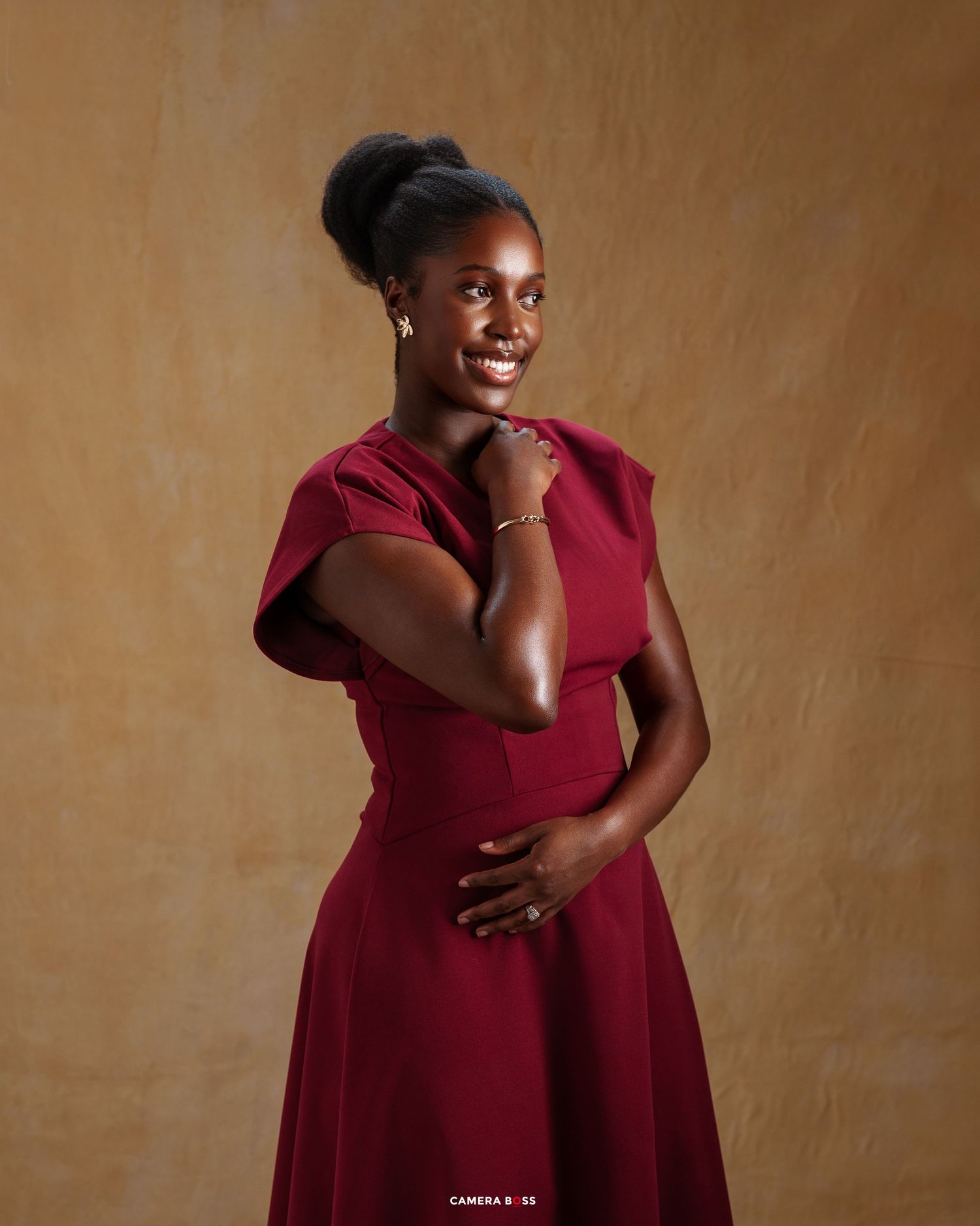 A person in a burgundy dress poses elegantly against a textured brown background with a warm smile.