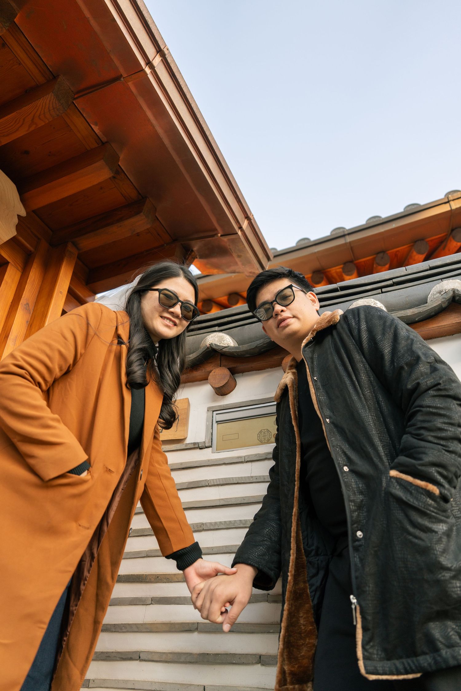 Two people in casual winter attire interact playfully on outdoor steps with wooden beams overhead.