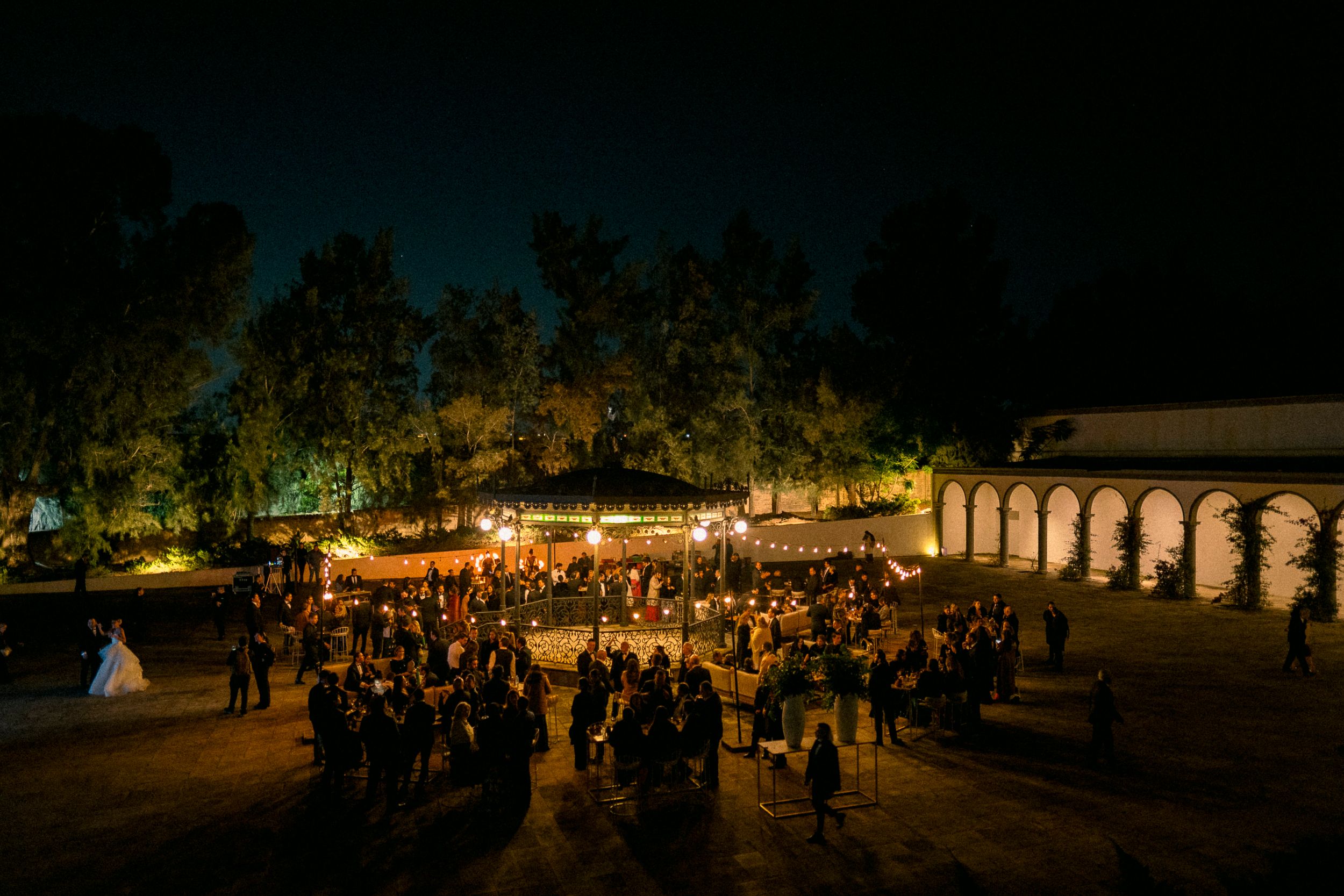 A nighttime outdoor event with glowing lights and crowds gathered under palm trees at a festival venue.