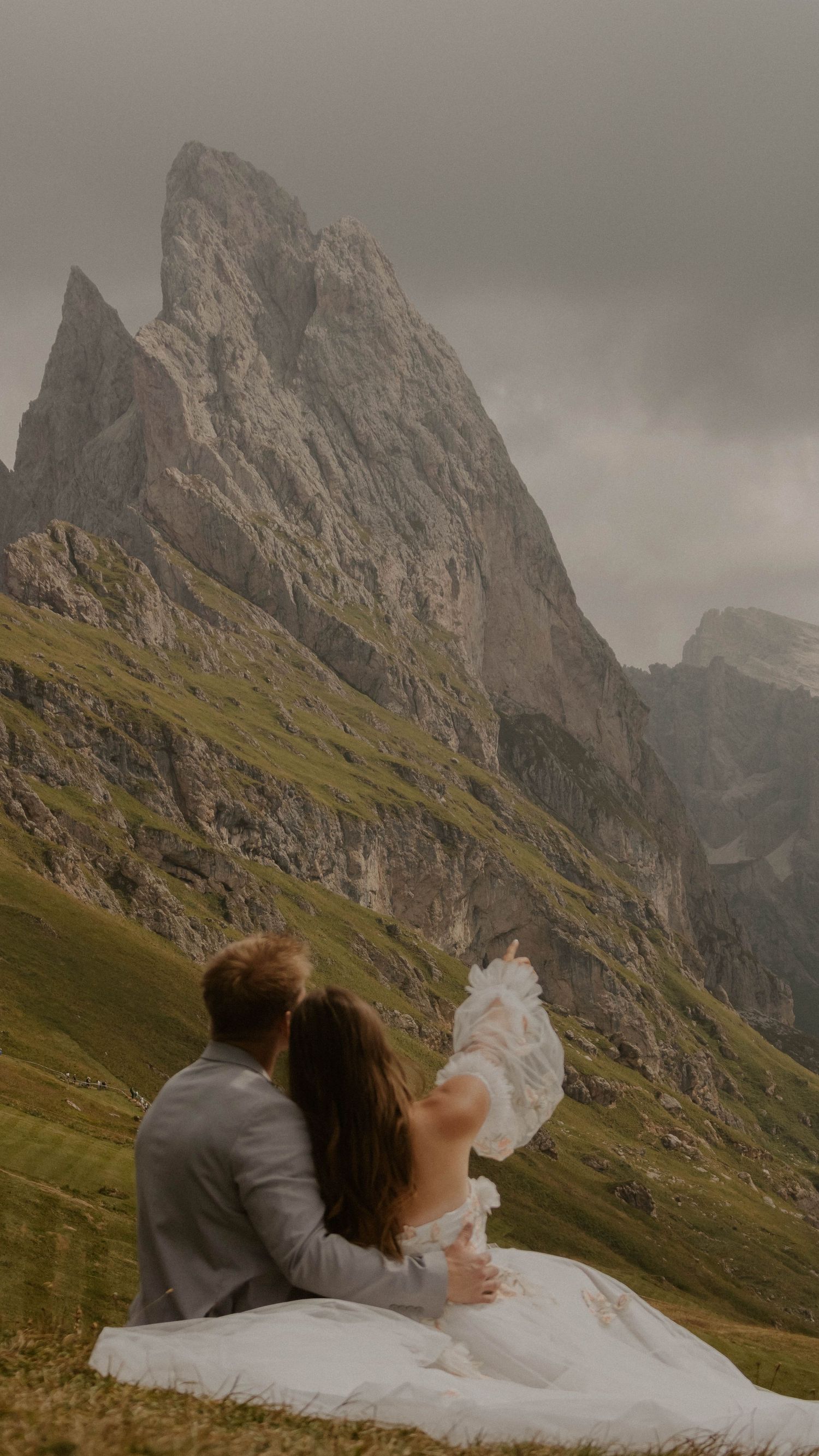 A couple in wedding attire sits on a grassy slope overlooking dramatic mountain peaks in the Alps on a cloudy day.