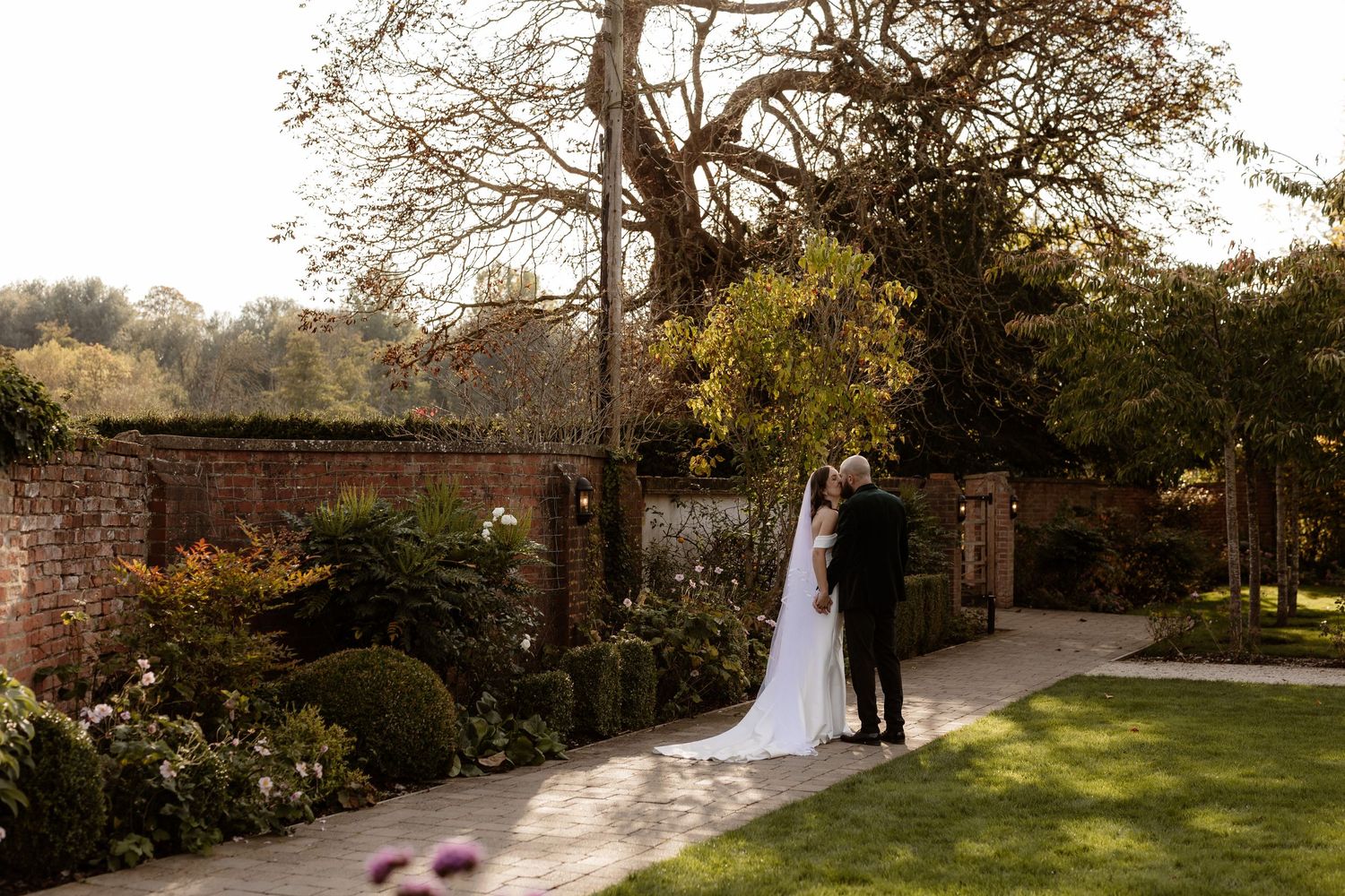 A romantic wedding couple poses in a sunlit garden setting during golden hour with stone architecture in the background.