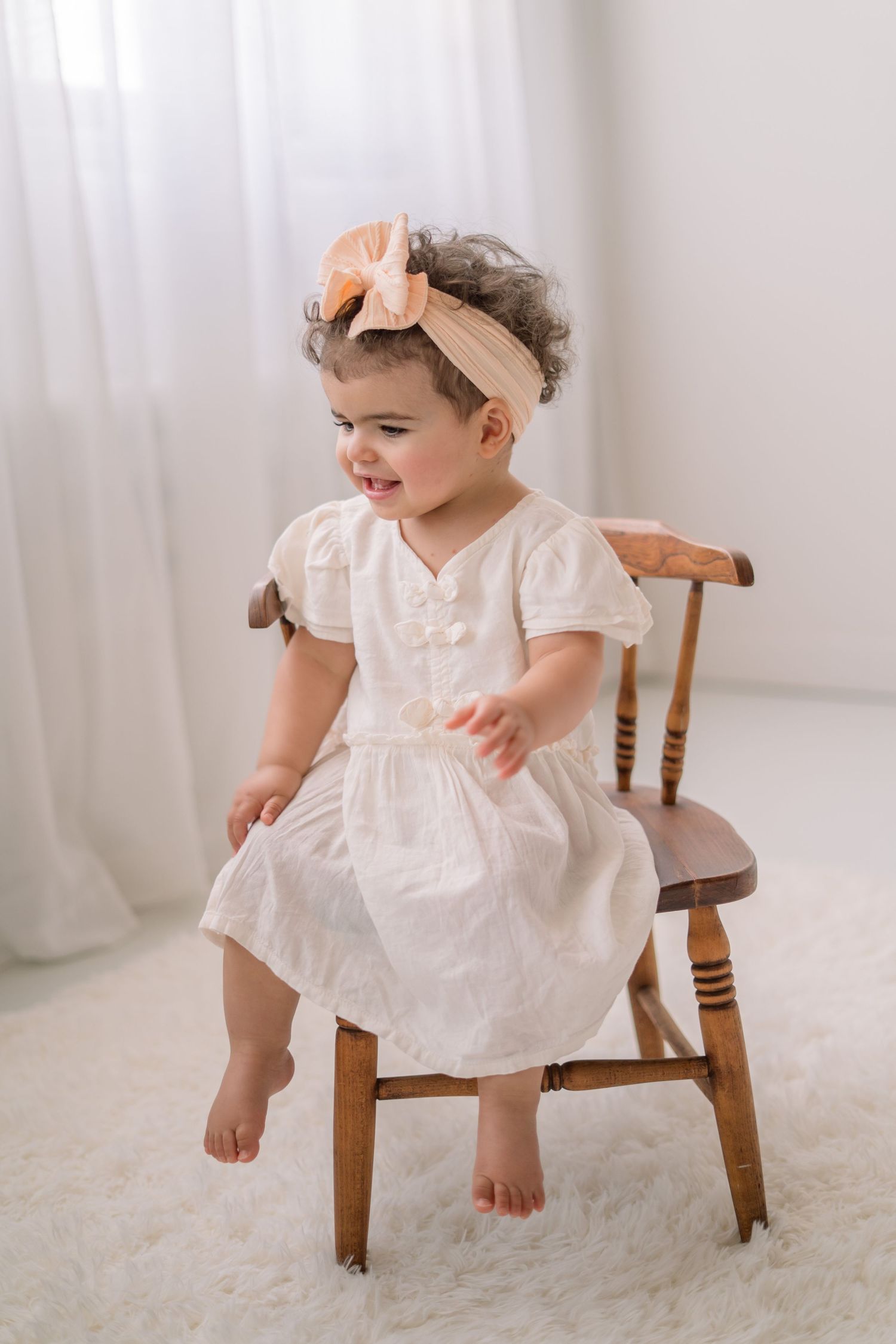 A small child in a white vintage dress sits and plays on a wooden chair against a light backdrop.