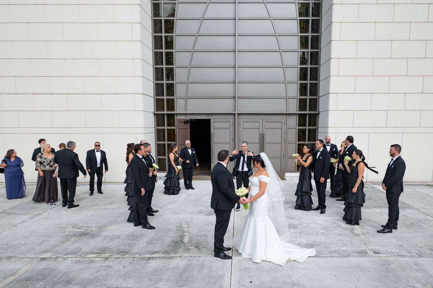 Wedding party poses together against a white stone building exterior during an elegant outdoor celebration.
