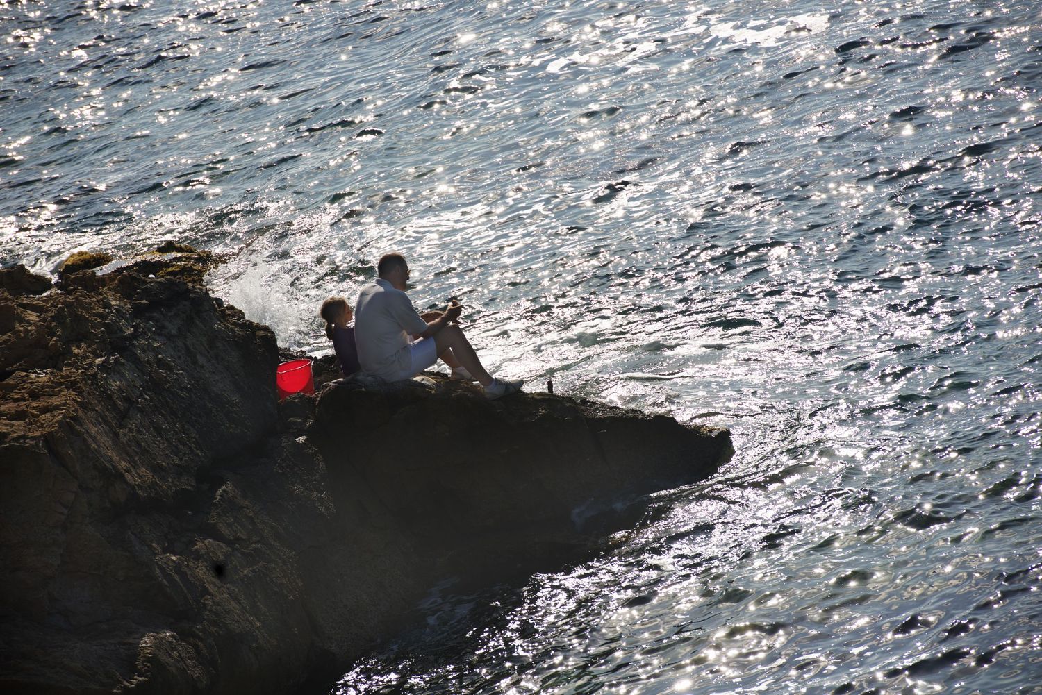 Person fishing on rocky cliff edge overlooking sparkling ocean.