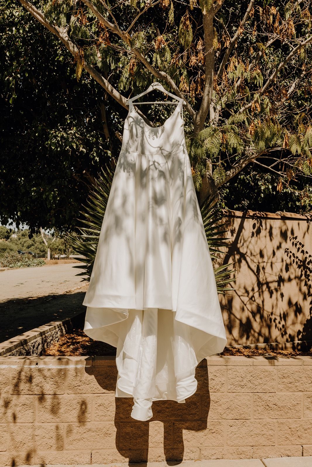 A white wedding dress hangs elegantly from a tree branch casting dappled shadows at River Garden Weddings and Events.