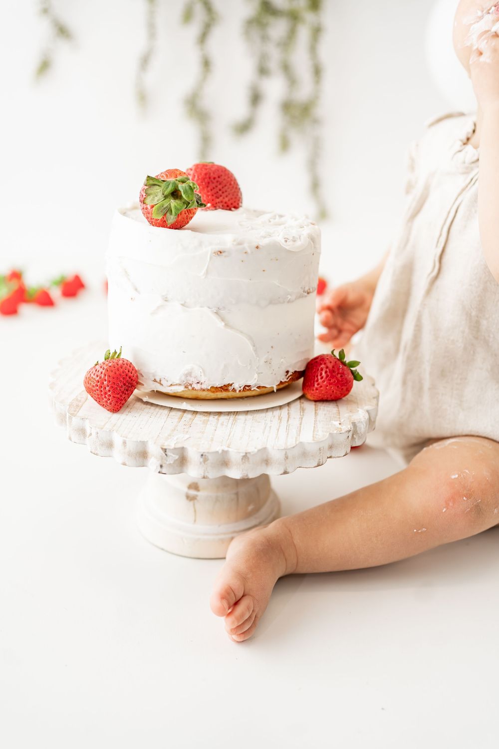 A white frosted cake decorated with fresh strawberries sits on an elegant cake stand against a light background.