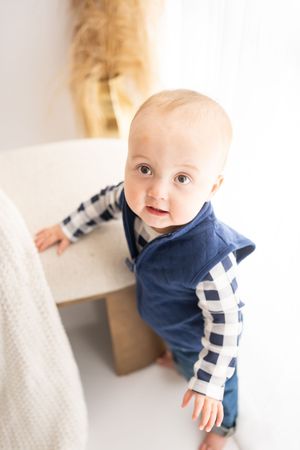Baby wearing navy and white striped sweater leans against white furniture in bright indoor setting.