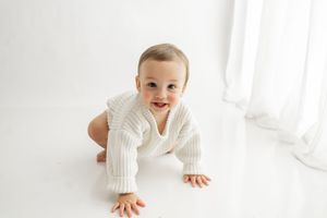 Baby in white onesie crawls and smiles on bright white background.