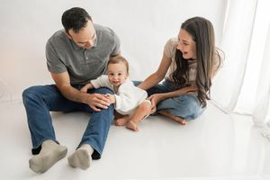 Family lying together on white floor in casual clothing during intimate lifestyle photo session.