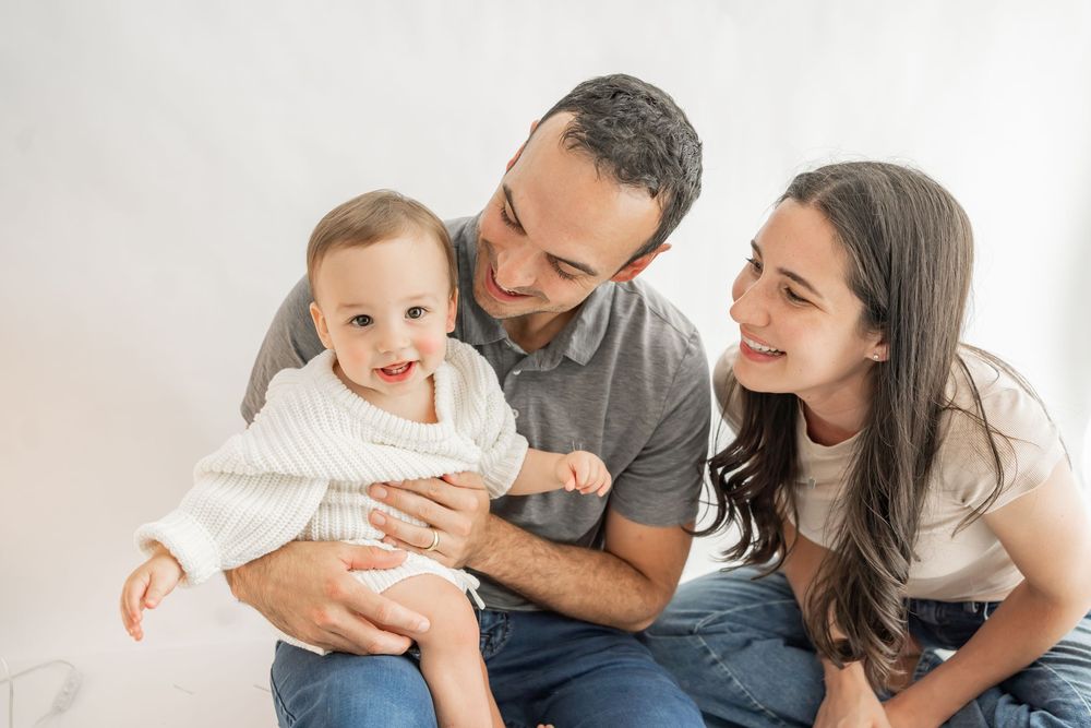 A family with a baby sits together on a white bed sharing tender moments and smiles.