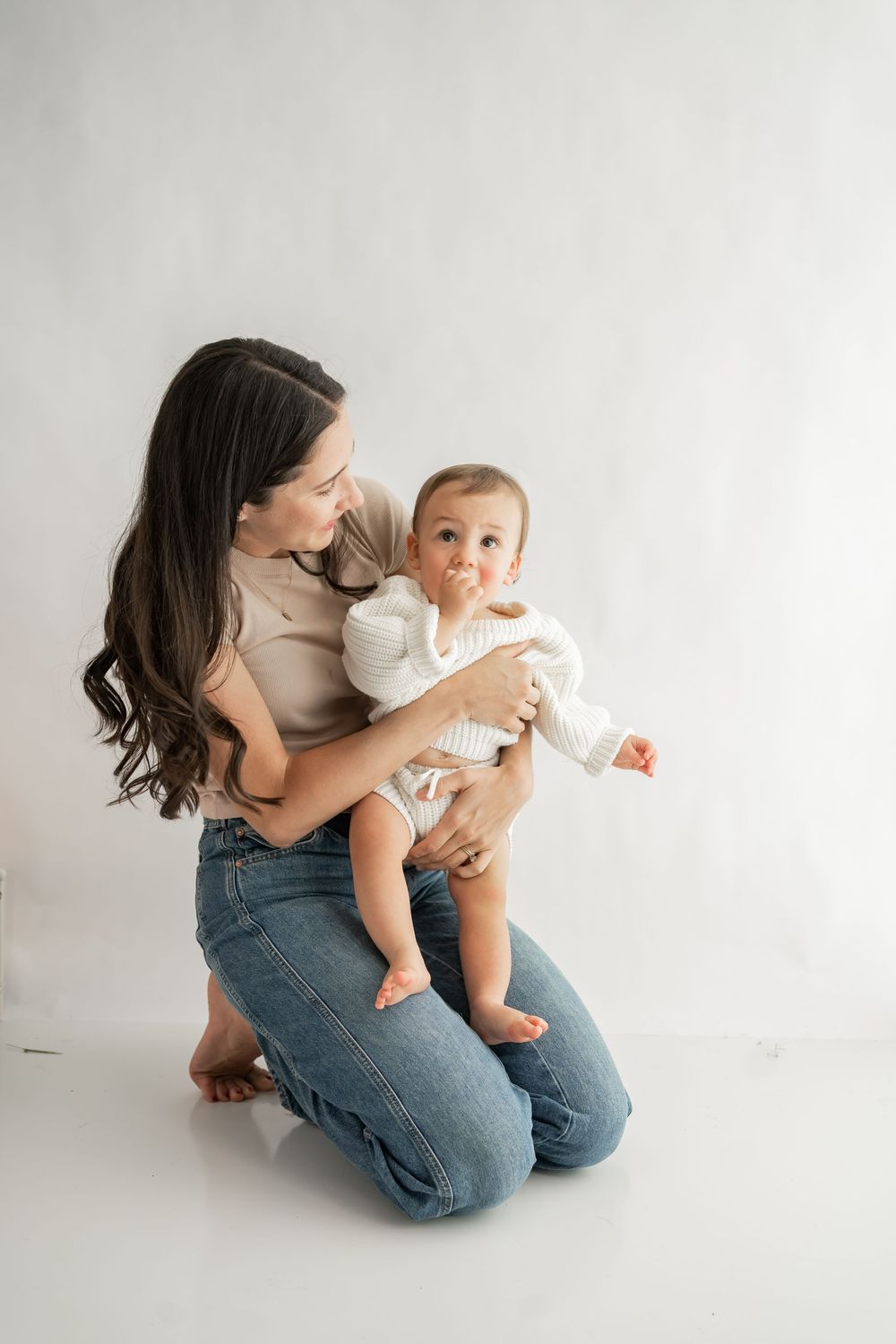 A parent in blue jeans kneels on a white floor while holding and interacting with a baby in a white onesie.