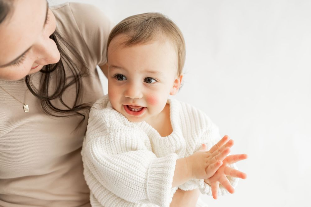 Baby in white sweater smiling and playing during indoor photoshoot with bright natural lighting.