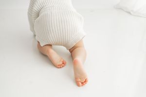 Baby wearing white knit romper crawls on white surface showing bare feet and legs.