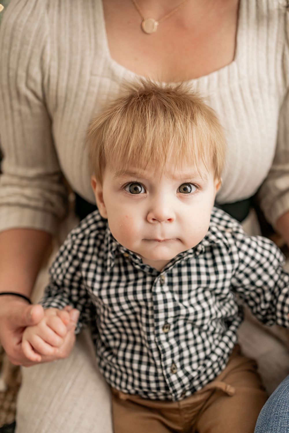 A baby wearing a black and white gingham shirt sits on a lap in a cozy indoor setting.