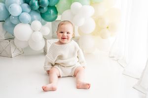 A small child in a cream sweater sits on white floor with teal and white balloon decorations in the background.