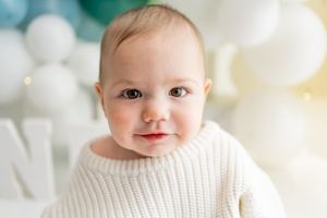 A small child wearing a cozy white knit sweater sits against a light background.