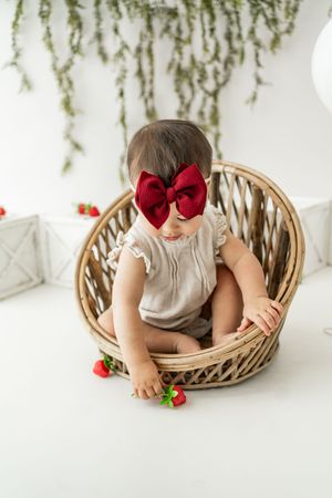 Baby wearing red bow and beige dress reaches for strawberries while sitting in woven basket against white backdrop.