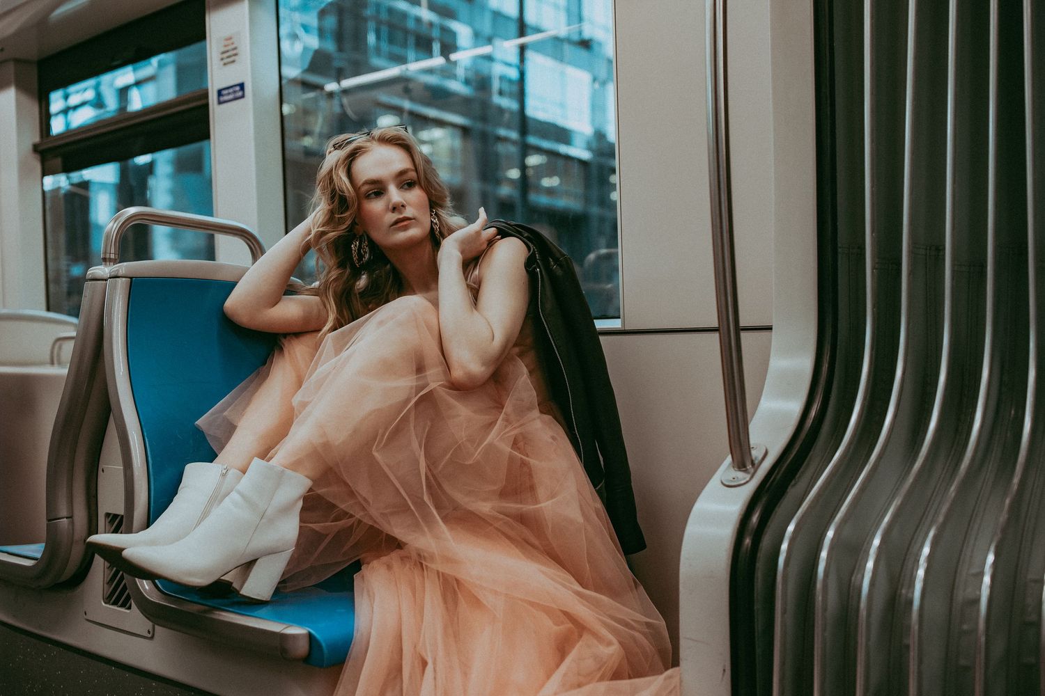 A person in a brown sweater and flowing peach gown poses on vintage turquoise seats of an empty train car.