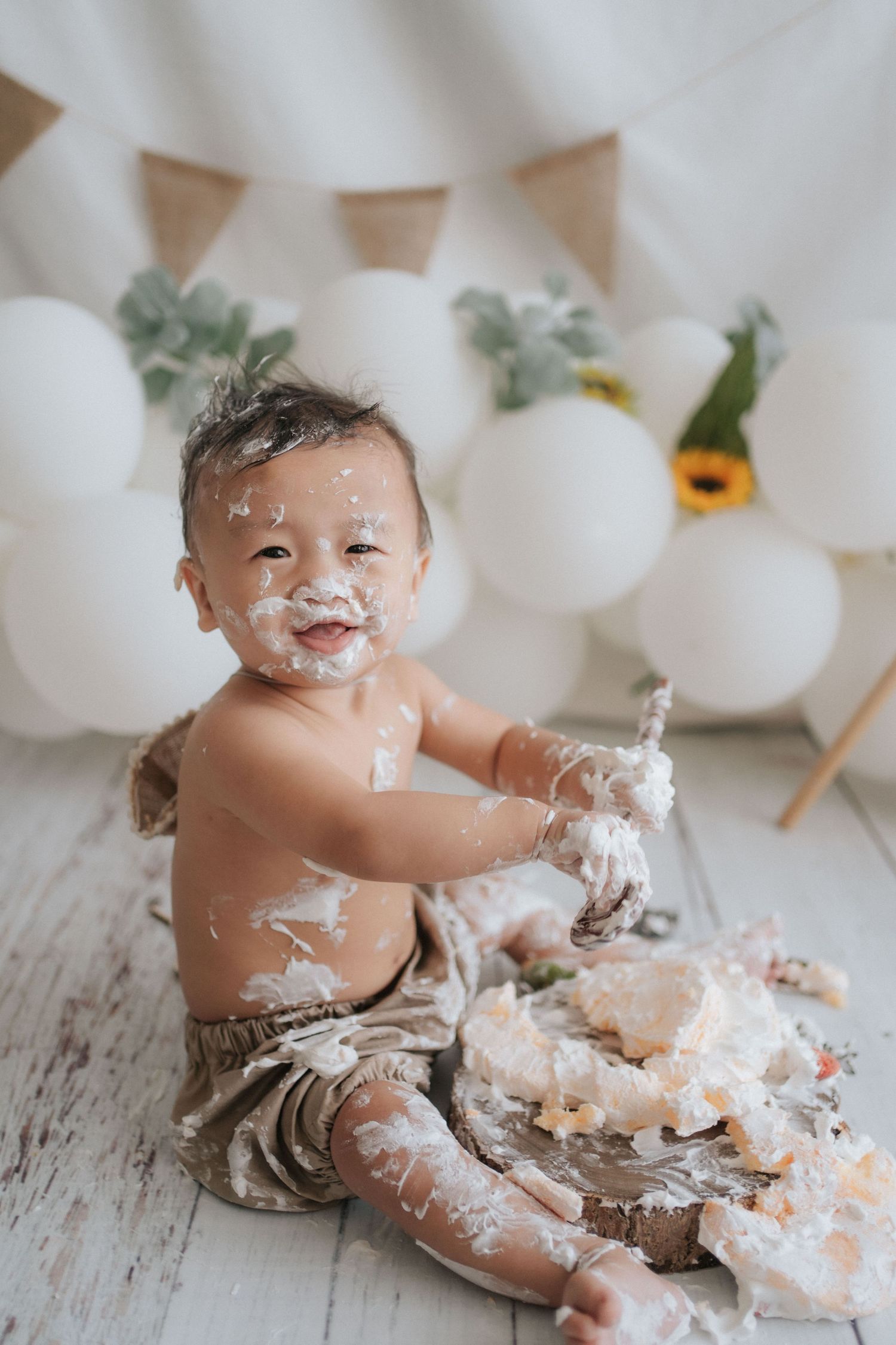 A messy cake smash photo session with white balloons and bunting decorations on a rustic wooden floor.
