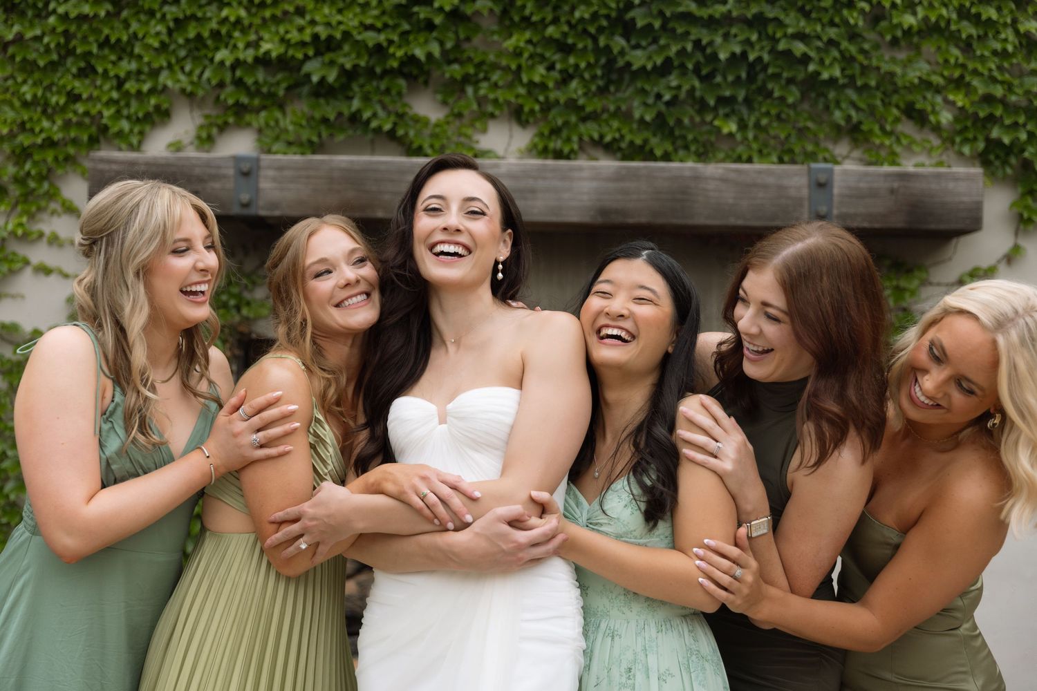 A group of women in formal attire laugh and embrace joyfully during a wedding celebration outdoors.