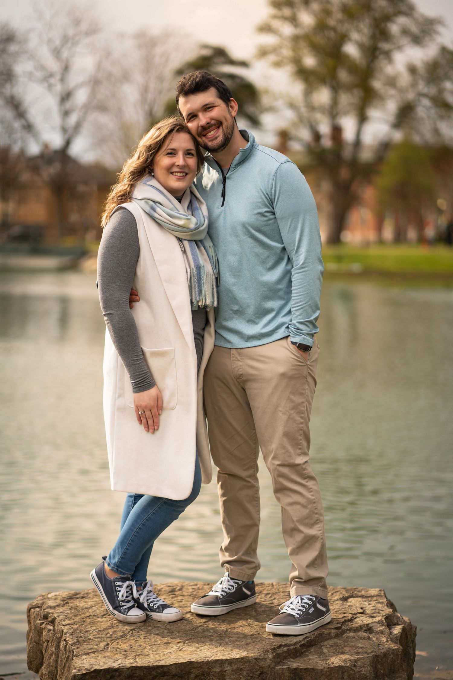 A couple stands together smiling on a rock by a lake wearing casual fall attire and matching sneakers.