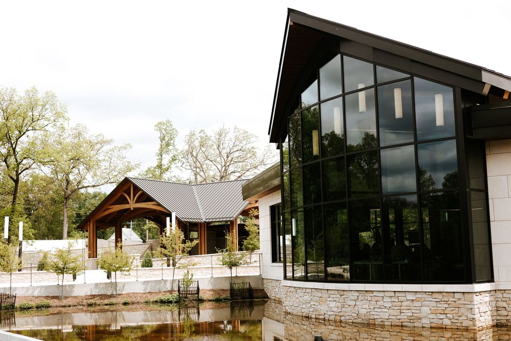 Modern building with large glass windows beside a tranquil pond, flanked by a wooden pavilion and lush trees.